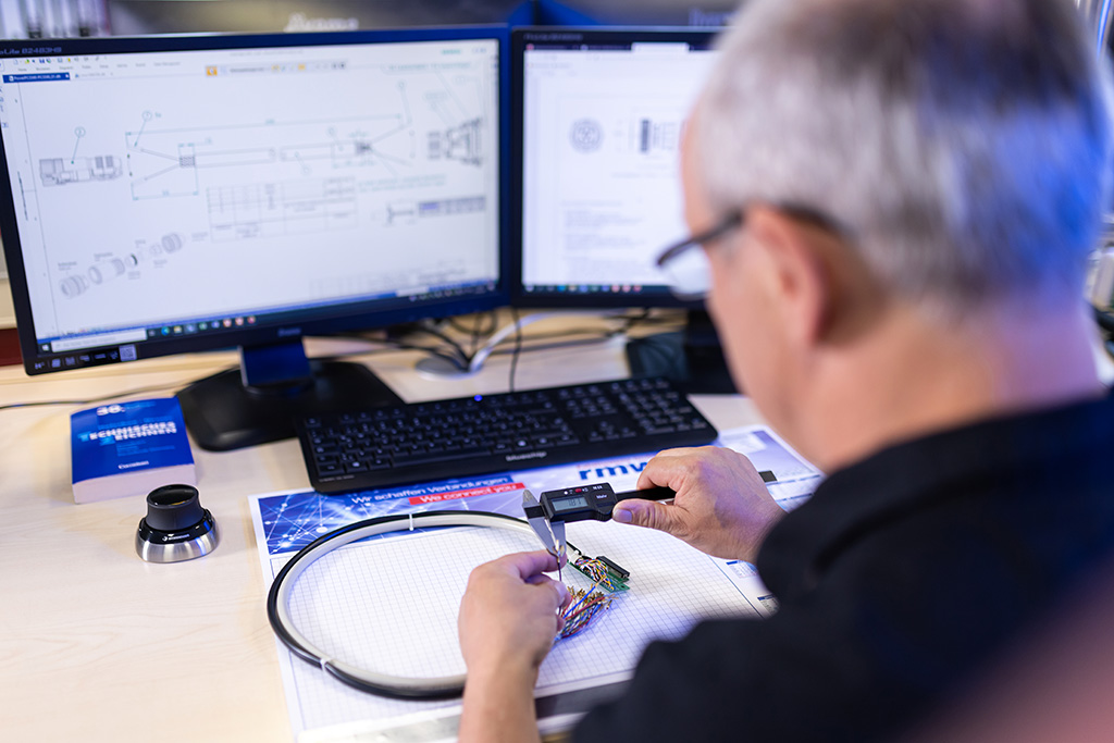 rmw-Mitarbeiter prüft Kabel mit Messschieber, im Hintergrund Monitor mit technischer Zeichnung